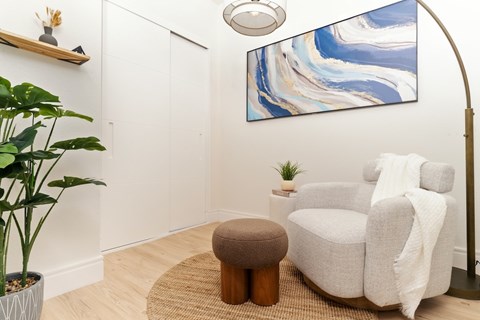 A living room with a white chair, a brown ottoman, and a large painting on the wall.
