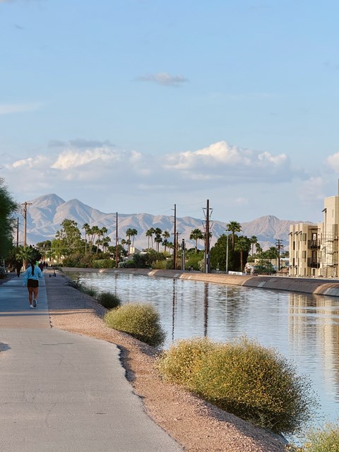 A person is walking on a path next to a body of water with mountains in the background.