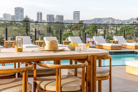A table with chairs and a hat on it is set up on a rooftop overlooking a pool and Los Angeles skyline.