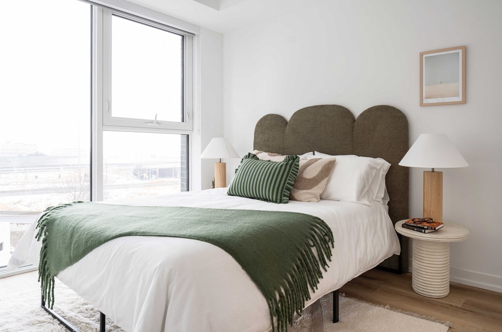 A bed with a white comforter and a green throw is in a room with a large window.