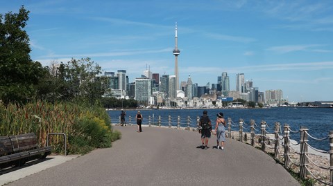 People walking on a path near a body of water with a city skyline in the background.
