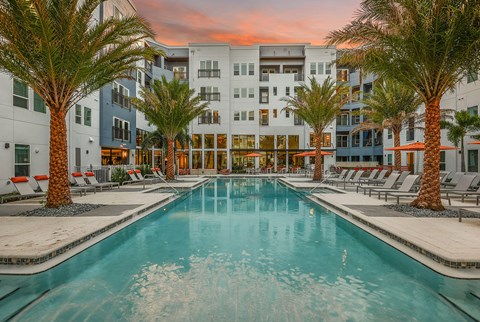 A swimming pool surrounded by palm trees and lounge chairs in front of a multi-story building.