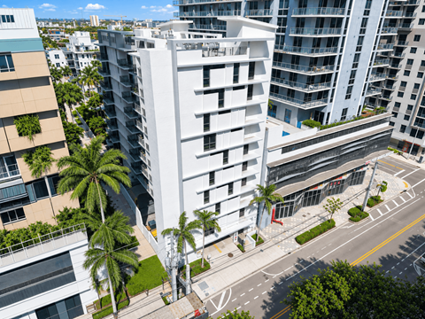 A white building with balconies is surrounded by other buildings and palm trees.
