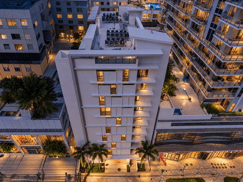 A modern white building with balconies is surrounded by other buildings and palm trees.