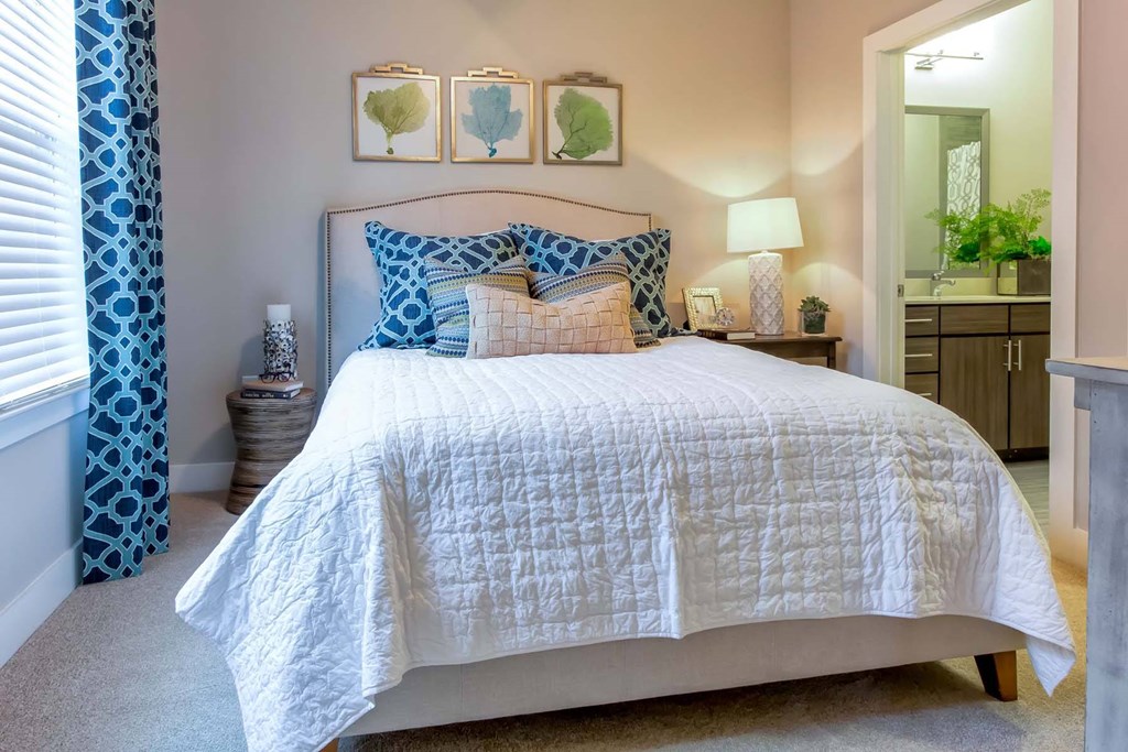 A bed with a white comforter and a blue pillow is in the center of the image at Wharf 7 Apartments, Charleston, 29492