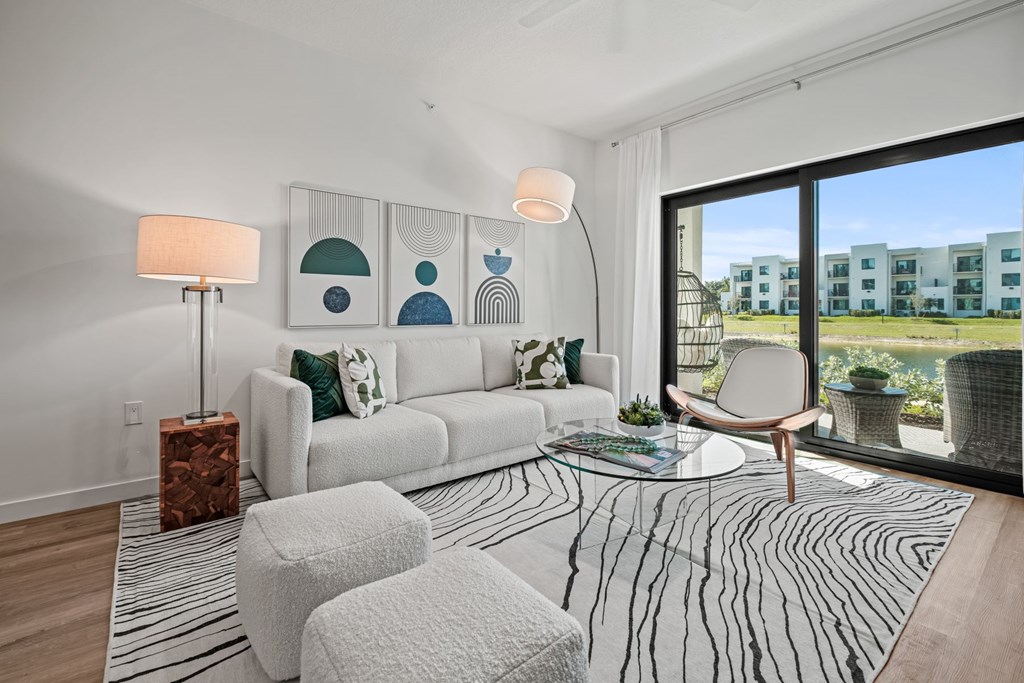 A modern living room with a white sofa, a wooden coffee table, and a large window with a view of the balcony overlooking the water
