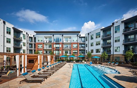 A large swimming pool surrounded by lounge chairs and umbrellas in front of apartment buildings.