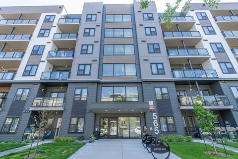 The image shows a modern apartment building with a grey and white exterior and a glass entrance.