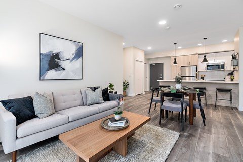 A modern living room with a grey sofa, a wooden coffee table, and a dining area in the background.