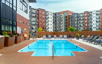 A large swimming pool surrounded by lounge chairs and apartment buildings.