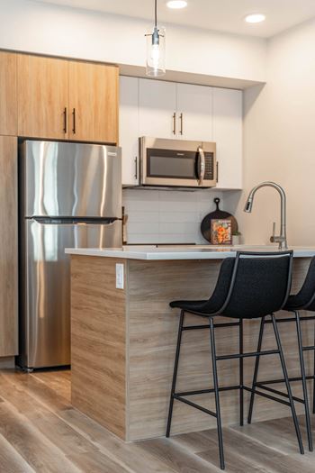 A kitchen with a stainless steel refrigerator, a microwave above the counter, and two black barstools.at Vibe Apartments at Arbour Lake, Calgary Alberta  