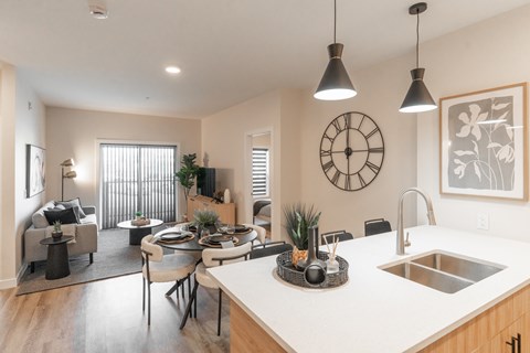 A modern kitchen with a dining table and chairs, a clock on the wall, and pendant lights hanging above the island.
