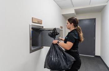 A woman is holding a black trash bag in front of a trash can.