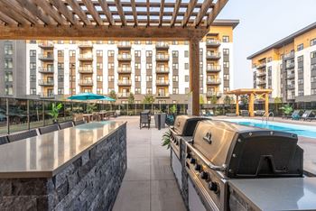 A large outdoor grill is in the foreground of a patio with a pool and apartment buildings in the background.