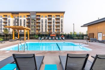 A large swimming pool with lounge chairs and a wooden pergola in front of a multi-story apartment building.
