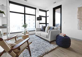 A living room with a grey couch, a wooden chair, a white rug, and a large window.at Raymond Block, Alberta