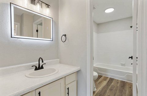 A white bathroom with a sink, mirror, and wooden floor at The Onyx Hoover Apartments, Alabama