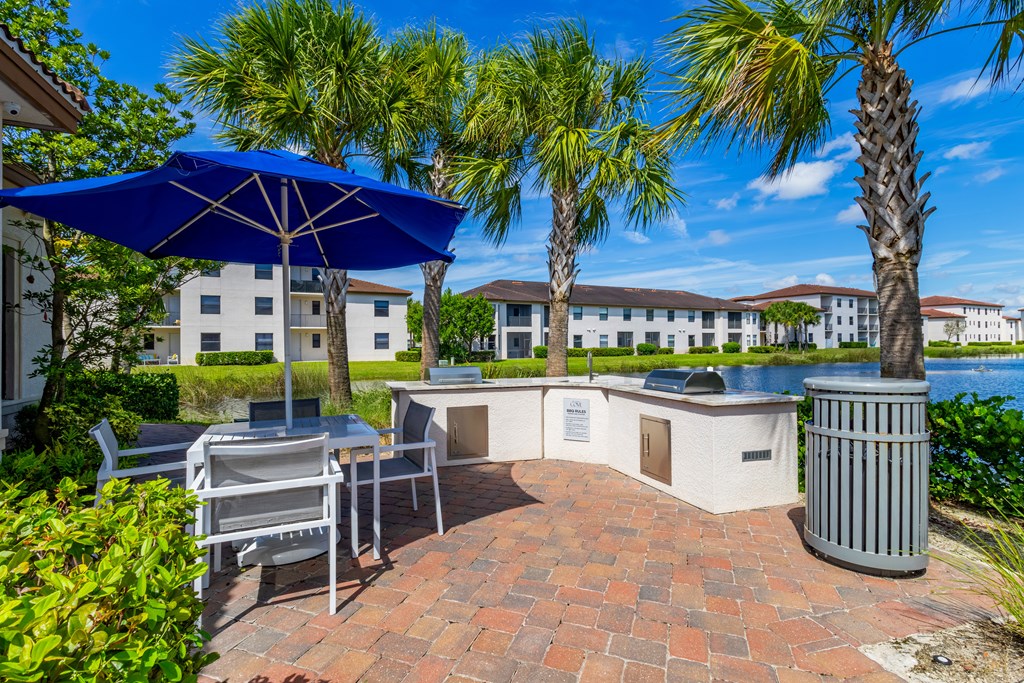 A patio with a table and chairs under an umbrella with a trash can and palm trees.