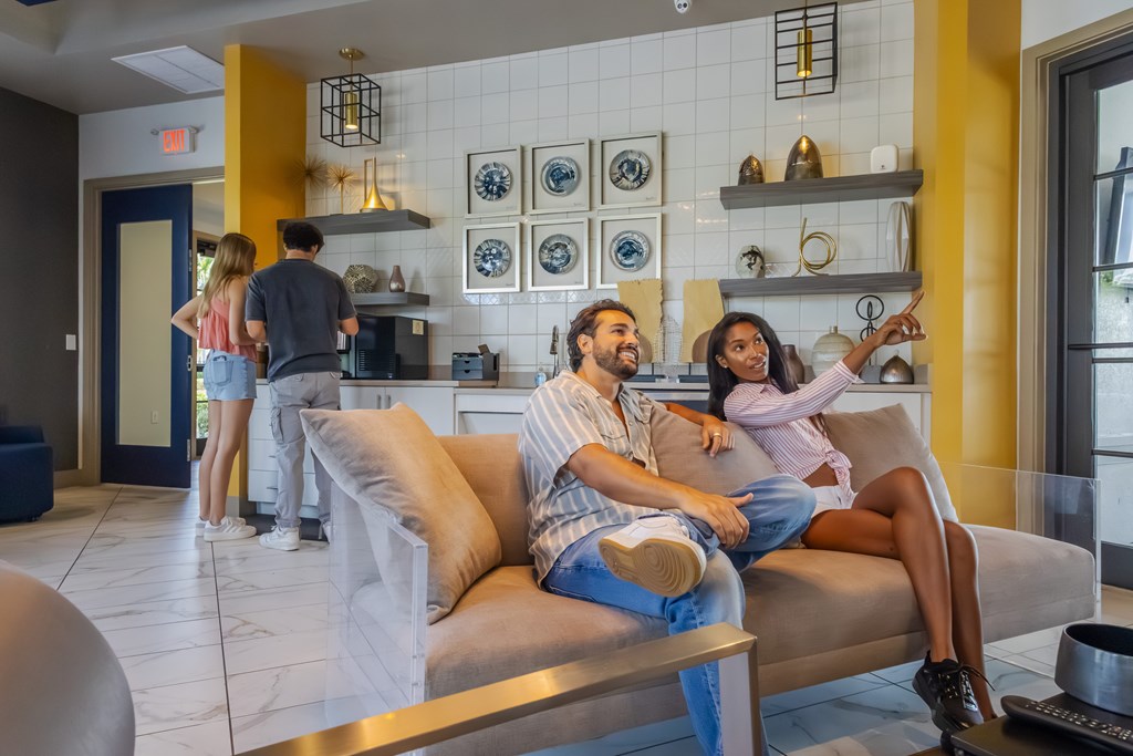 A couple is sitting on a couch in a brightly lit room with a kitchen area in the background.