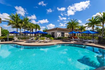 A swimming pool surrounded by palm trees and lounge chairs under blue umbrellas.