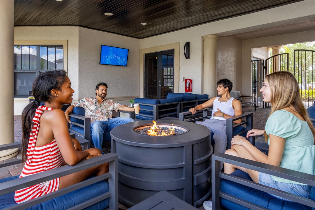 A group of people are sitting around a fire pit in a room with a TV screen on the wall.
