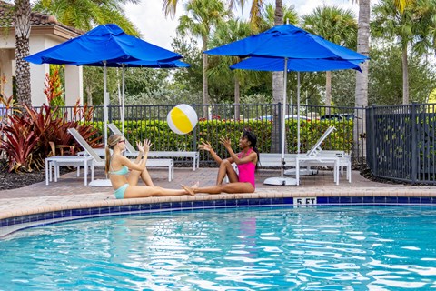Two women sunbathing by a pool with blue umbrellas.
