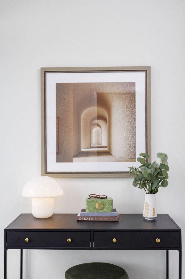a framed picture on a wall above a table with a lamp and a vase