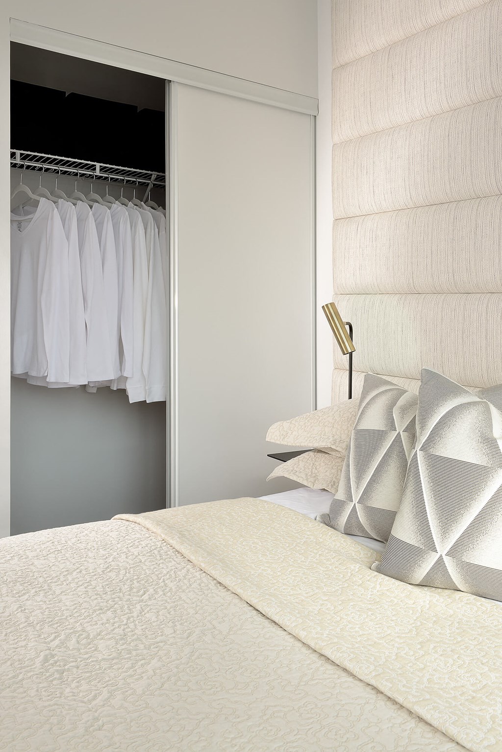 A bed with a white comforter and pillows with a white closet with clothes hanging in the background.