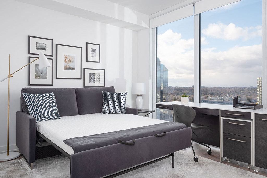 A bedroom with a large bed, a desk with a chair, and a view of the city through the window.