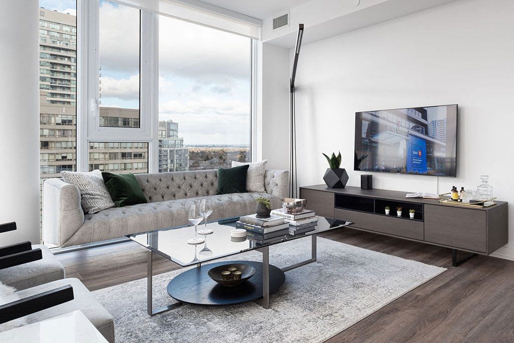 A modern living room with a grey sofa, a coffee table, and a large window overlooking the city.