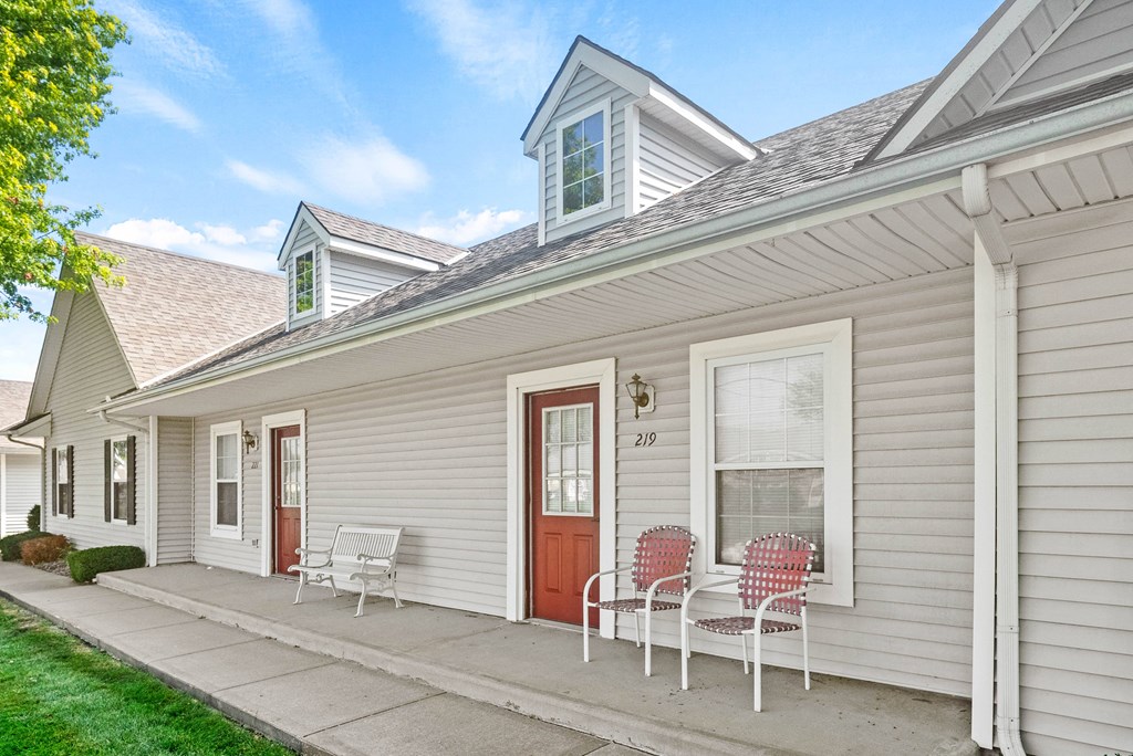 the front porch of a white house with two chairs and a red door