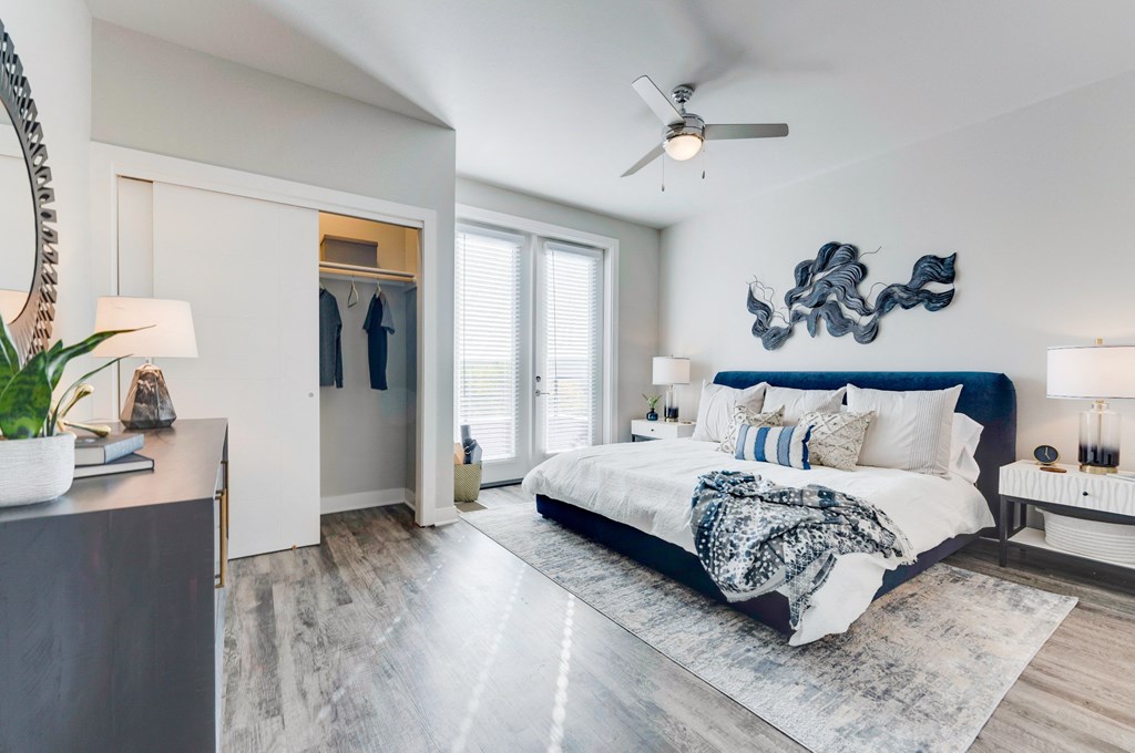 Apartment bedroom with wood floor, large bed with headboard, floor to ceiling windows and open closet.