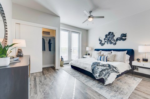 Apartment bedroom with wood floor, large bed with headboard, floor to ceiling windows and open closet.