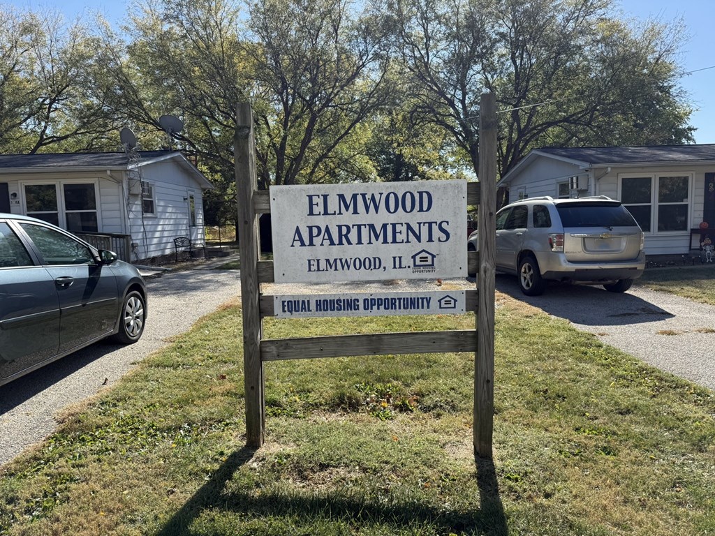 A sign for Elmwood Apartments stands in front of a grassy area with cars and houses in the background.