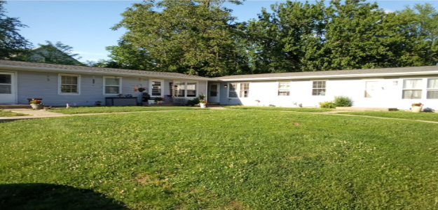 A grassy area in front of a white building with trees in the background.