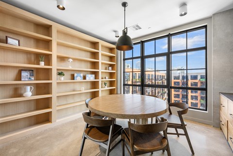 A dining room with a round table and chairs, a shelf with decorative items, and a view of the city through large windows.