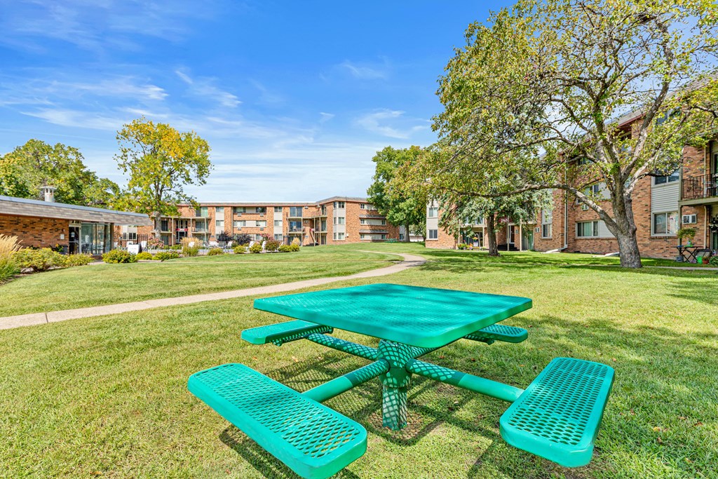 A green picnic table and benches are set up on a grassy area in front of a brick building.