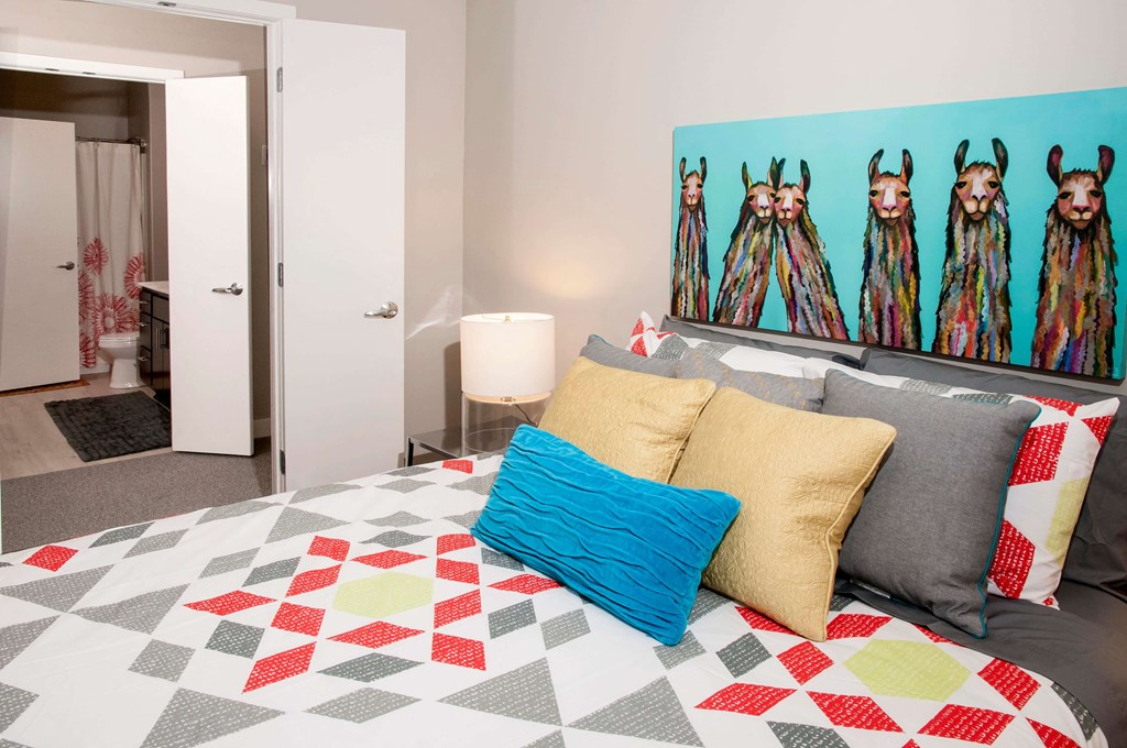 Master bedroom leading into walk through closet and private bathroom. Bed is staged with a brightly colored comforter and a large piece of art hangs above the headboard.