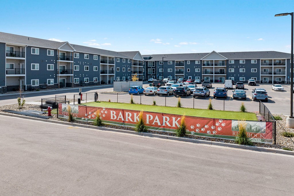A parking lot in front of apartment buildings with a sign that says Bark Park. at Red Rock Apartments, South Dakota