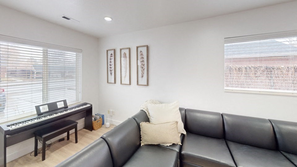 A black leather couch sits in a living room with a piano and two framed pictures on the wall.