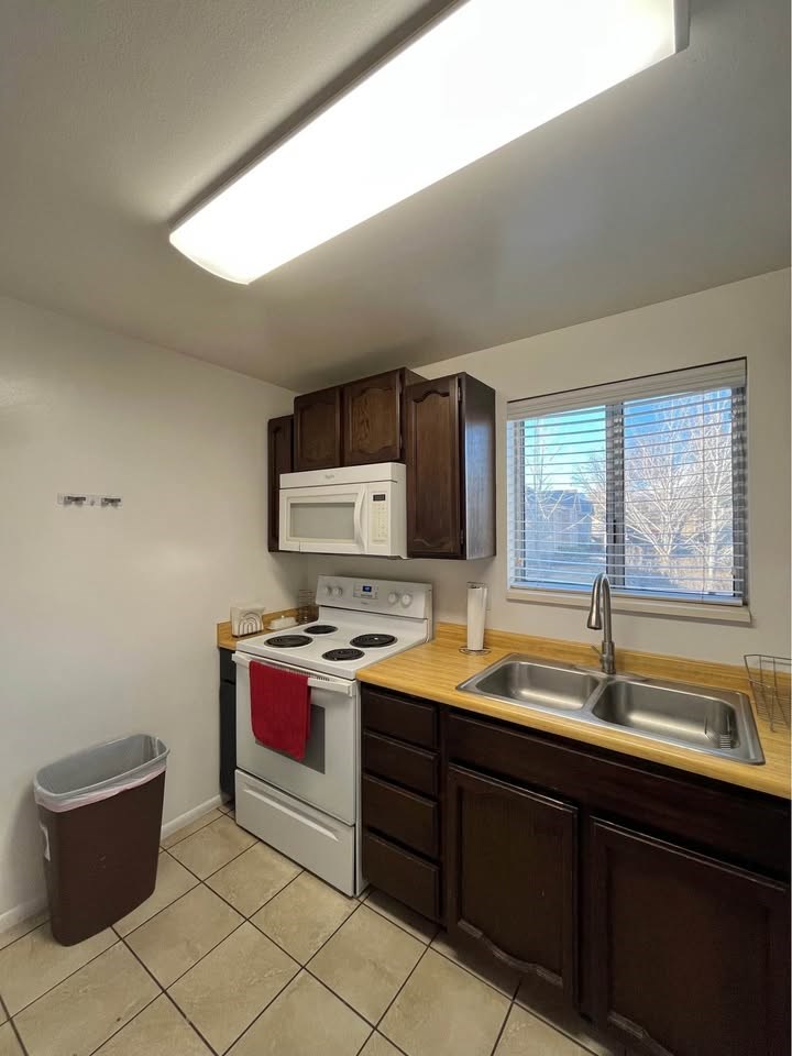 A kitchen with a white microwave, a white oven, a brown trash can, brown cabinets, a brown countertop, and a brown sink.