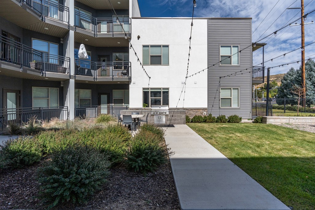 an exterior view of an apartment building with a sidewalk and grass at The Flats at Riverwoods, Provo, Utah