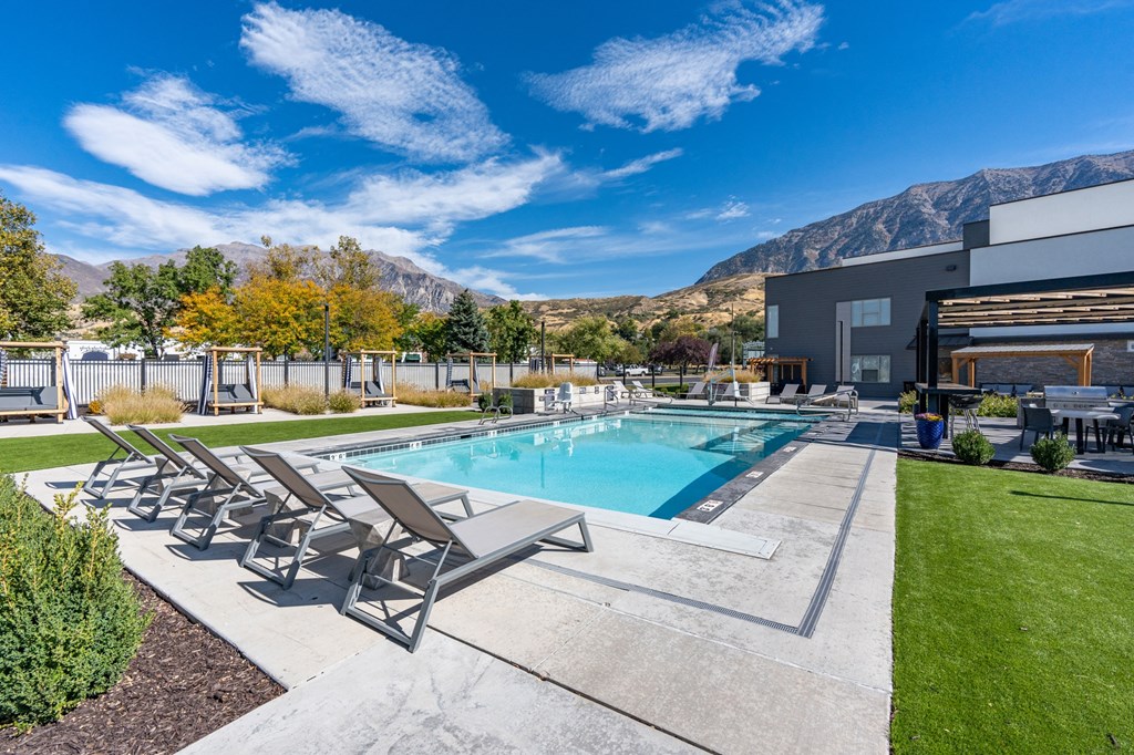 a swimming pool with patio furniture and a building with mountains in the background at The Flats at Riverwoods, Provo, UT