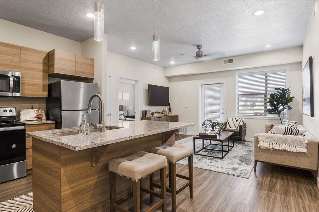 a kitchen with an island and stools in front of a living room with a couch and at The Flats at Riverwoods, Provo, 84604