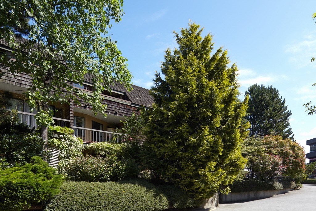 a building with a gray roof and green bushes and trees in front of it