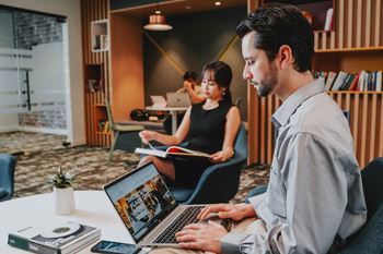 A man is working on a laptop while a woman reads a book in the background.