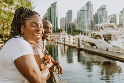 Two people are standing by the water with boats and buildings in the background.