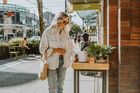 A woman in a white jacket and ripped jeans stands on a sidewalk next to a table with a potted plant on it.
