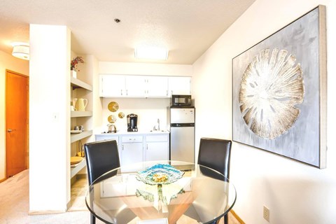 A dining room with a glass table and chairs, a framed artwork on the wall, and a kitchen area in the background.