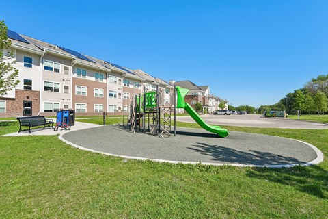 A playground with a green slide and a bench in front of apartment buildings.
